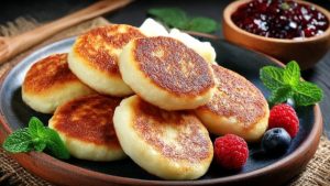 Домашні сирники з рум’яною скоринкою, подані з ягодами, сметаною та варенням